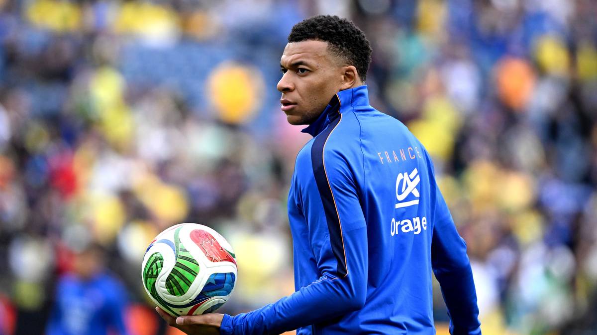 Piłkarz w niebieskiej koszulce trzyma piłkę w dłoni na tle stadionu z kibicami.