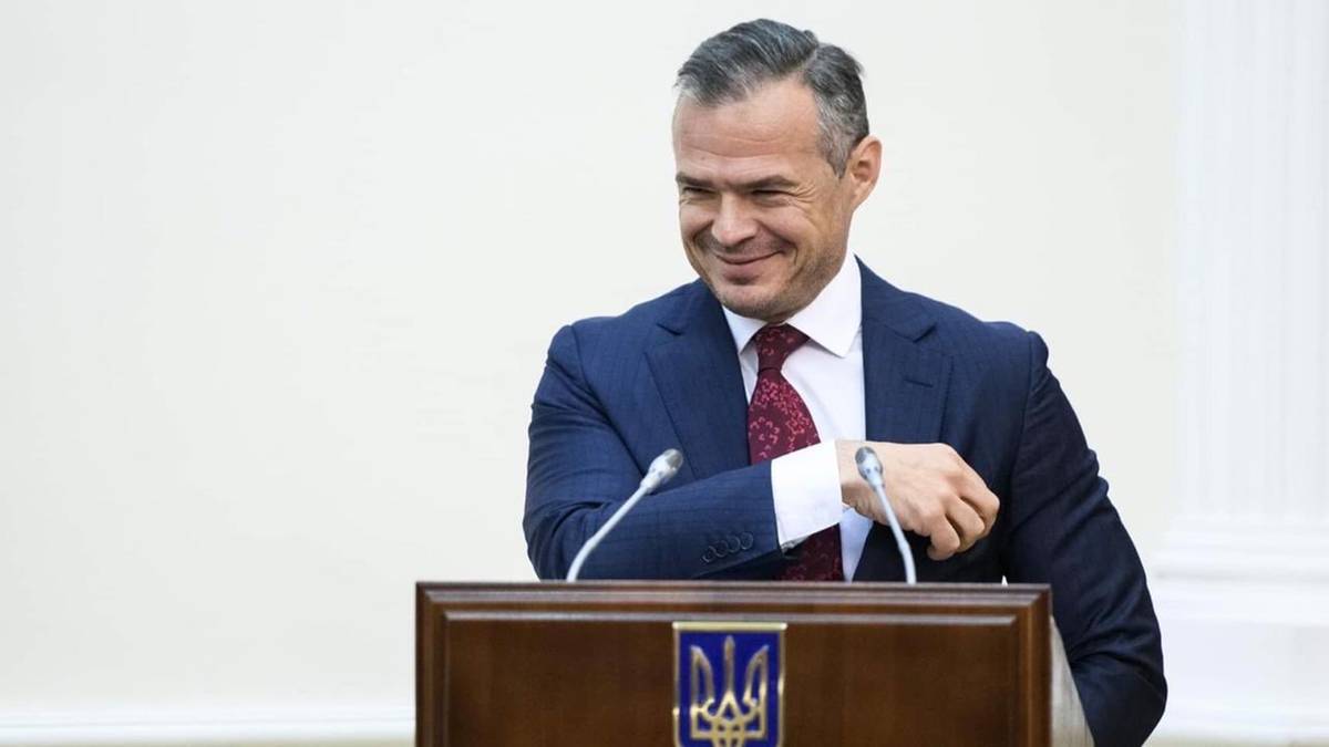 Mężczyzna w garniturze i krawacie uśmiecha się za m&oacute;wnicą.