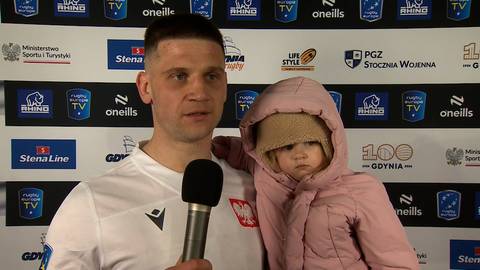 Wojciech Piotrowicz trzyma w ramionach małe dziecko ubrane w r&oacute;żowy kombinezon i czapkę, trzymając mikrofon. Tło stanowi ściana z logotypami sponsor&oacute;w.