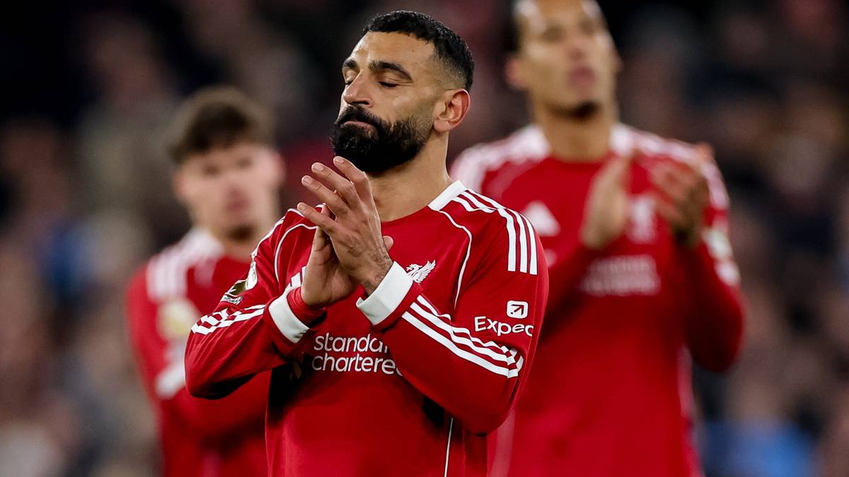 Piłkarz Liverpoolu z zamkniętymi oczami i złożonymi dłońmi w geście modlitwy lub refleksji, w tle inni zawodnicy w czerwonych strojach.