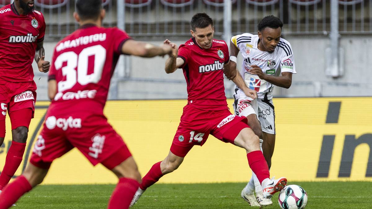 Piłkarz w czerwonym stroju pr&oacute;buje odebrać piłkę zawodnikowi w białej koszulce.