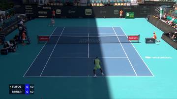 Dwóch tenisistów na korcie tenisowym podczas meczu. Jeden gracz w zielonej koszulce stoi blisko siatki, a drugi w czerwonej koszulce znajduje się na końcu kortu. Na ekranie widoczny jest wynik meczu: Tiafoe 0, Sinner 0.