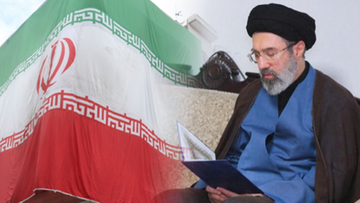 Mężczyzna w czarnym turbanie i okularach siedzi, czytając dokument, obok rozwieszonej flagi Iranu.