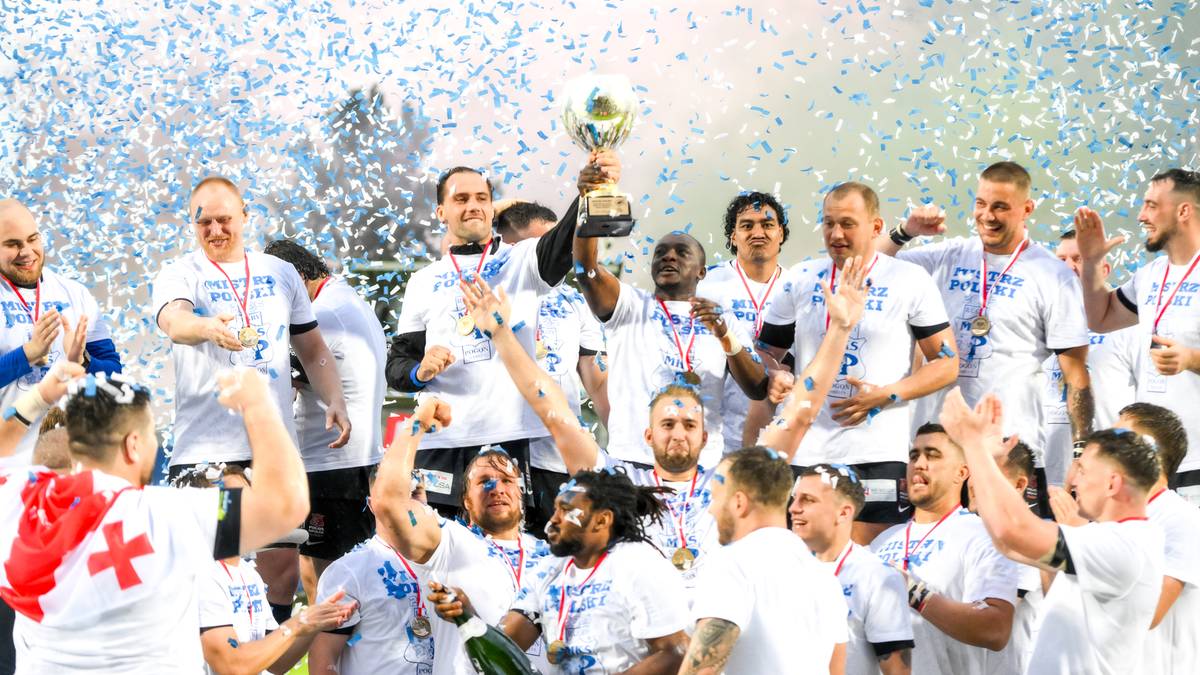 Drużyna rugby celebruje zdobycie pucharu, unosząc go nad głowami w otoczeniu konfetti i rozlewającego się szampana.
