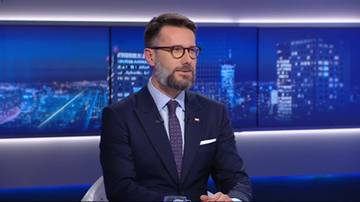 Mężczyzna w garniturze i okularach siedzi, mówiąc. Tło to ekran z widokiem nocnego miasta.