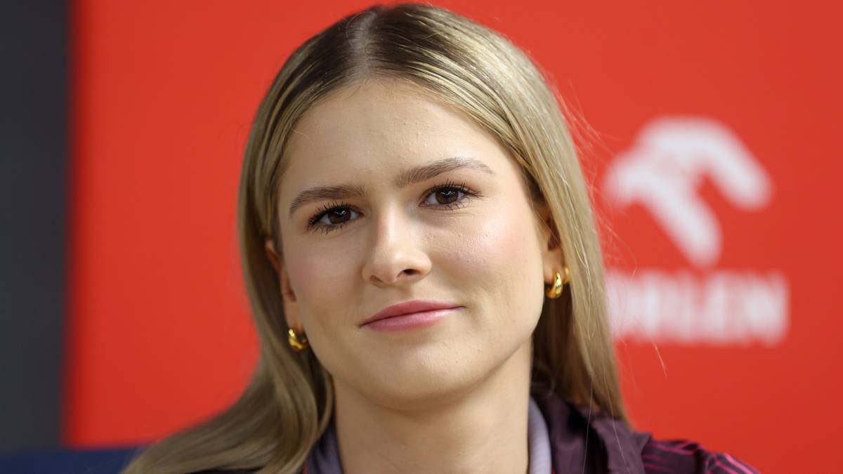 Portret kobiety z blond włosami, lekko uśmiechnięta, z widocznym logo sponsora w tle.