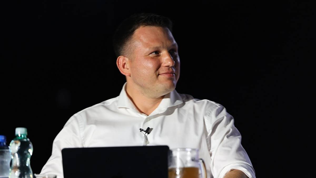 Mężczyzna w białej koszuli siedzi przy stole z laptopem i piwem na tle ciemnego tła.