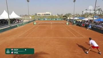 Gracz w białej koszulce i czerwonych spodenkach gra w tenisa na ceglanej nawierzchni kortu. W tle widoczna jest siatka, trybuny z widzami oraz namioty.