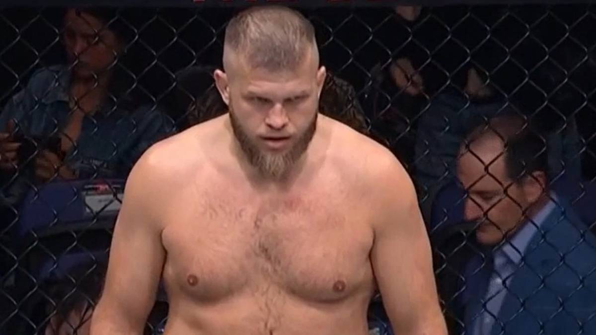 Marcin Tybura, polski zawodnik MMA, z brodą i krótko ostrzyżonymi włosami, stoi w klatce przed walką, jego twarz wyraża skupienie.