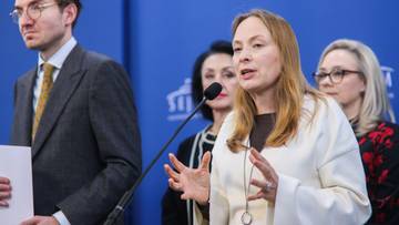 Kobieta w białej bluzce przemawia, gestykulując rękami, w tle mężczyzna w garniturze.