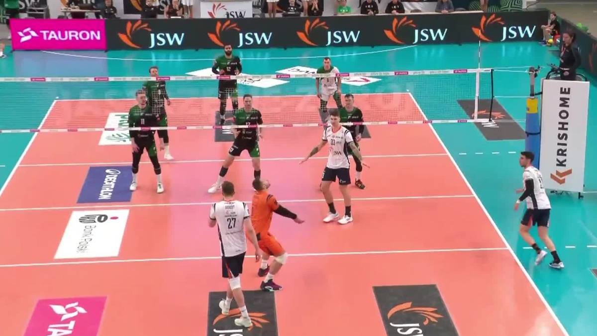 Mecz siatkówki halowej pomiędzy drużyną w zielonych strojach a drużyną w białych strojach, z widocznym zawodnikiem wykonującym atak.