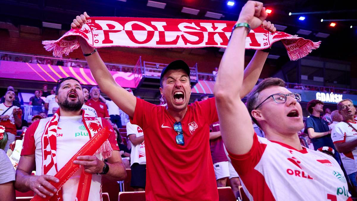 Trzech mężczyzn kibicuje podczas meczu siatkówki, trzymając flagę z napisem "POLSKA" i unosząc ręce w geście radości.