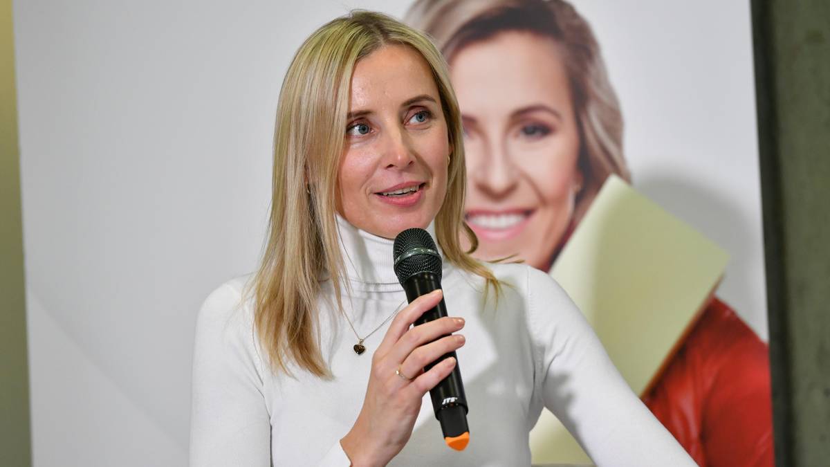 Kobieta w białym golfie trzyma mikrofon i mówi. Za nią znajduje się plakat z uśmiechniętą kobietą.