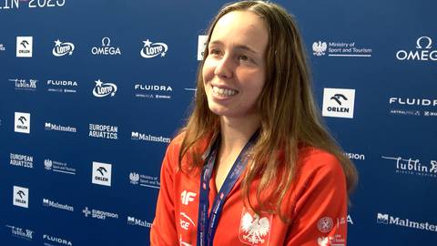 Uśmiechnięta młoda kobieta w czerwonej bluzie z kapturem, z medalem na szyi, na tle niebieskiej ściany z logo.