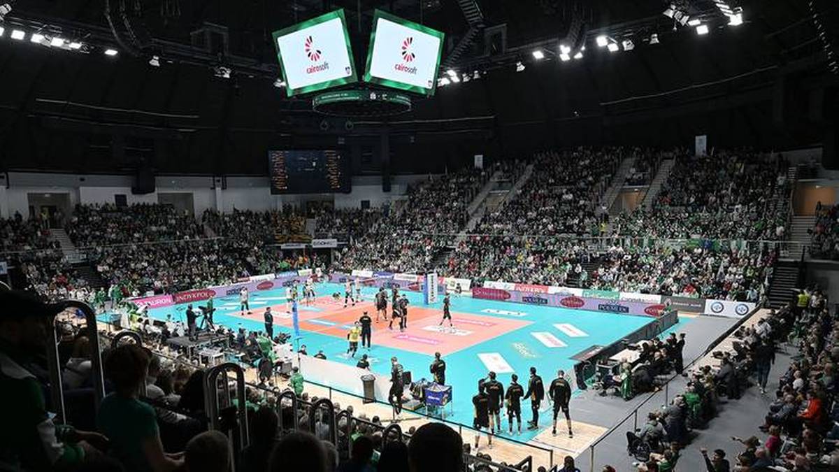 Widok z trybun na halę sportową podczas meczu siatkówki. Na niebieskim boisku z białymi liniami rozgrywany jest mecz, wokół boiska znajduje się duża widownia wypełniona kibicami. Nad boiskiem wiszą dwa ekrany.