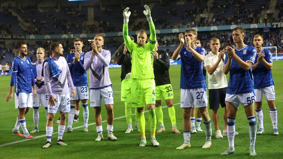 Drużyna piłkarska Lecha Poznań w zielonych strojach treningowych, z bramkarzem w jaskrawozielonym stroju, pozdrawia kibic&oacute;w na boisku piłkarskim.