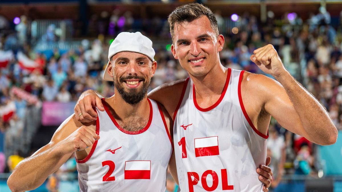 Dwóch mężczyzn w strojach sportowych z polskimi flagami na piersiach, uśmiechnięci, pozują do zdjęcia z ramionami na sobie.