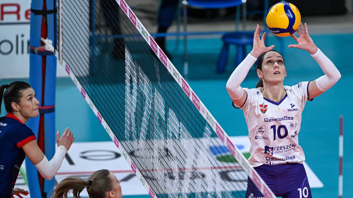 Siatkarka z numerem 10 na koszulce, w białej koszulce i granatowych spodenkach, podrzuca piłkę do zagrywki na tle siatki i innej siatkarki.