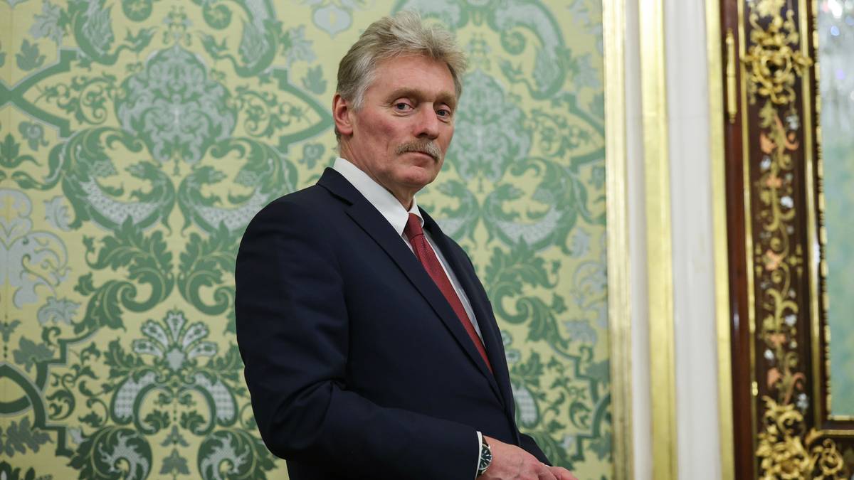 Dmitrij Pieskow w garniturze i krawacie stoi na tle ozdobnej tapety.