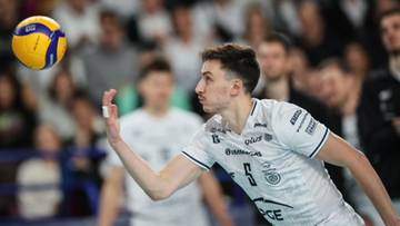 Siatkarz w białej koszulce z numerem 5 zagrywa piłkę do przodu.