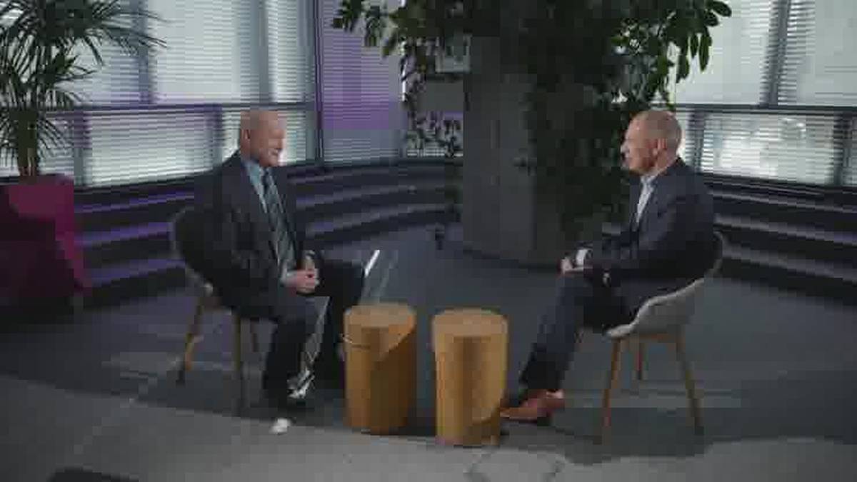 Dw&oacute;ch mężczyzn w garniturach siedzi naprzeciwko siebie na krzesłach w studiu nagraniowym. Pomiędzy nimi znajdują się dwa ż&oacute;łte pufy. W tle widać rośliny i nowoczesne okna.