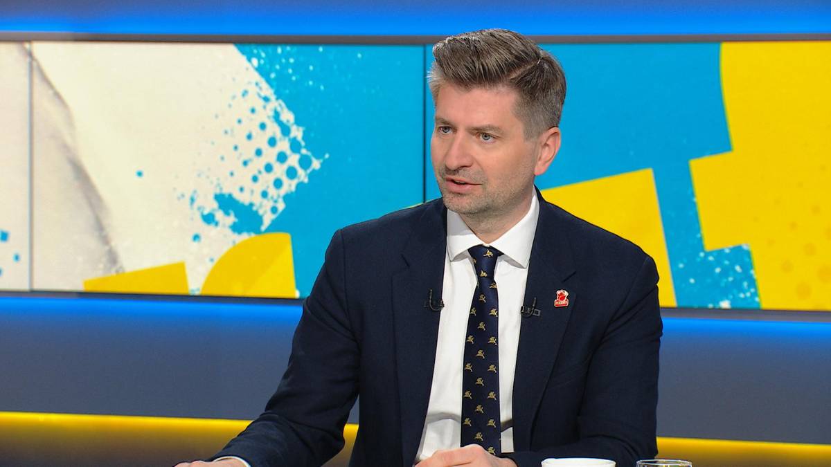 Krzysztof Śmiszek, europoseł, w ciemnym garniturze i białej koszuli z krawatem, siedzi podczas wywiadu telewizyjnego.