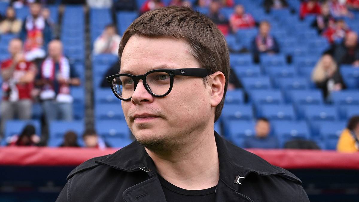 Mężczyzna w okularach i czarnej kurtce stoi na tle trybun stadionu.