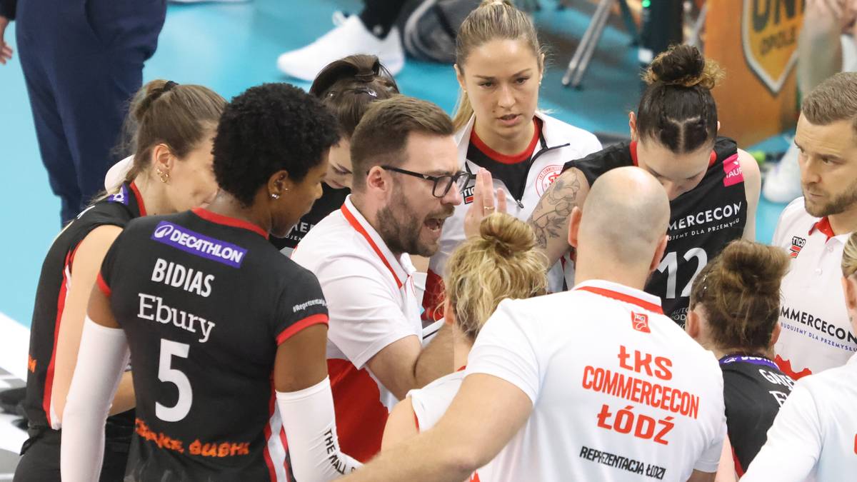 Grupa zawodniczek siatkarskich w czarnych strojach z numerami i nazwiskami, skupiona wokół trenera w białej koszulce, podczas narady taktycznej.