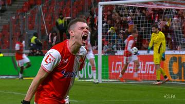 Piłkarz w czerwonej koszulce z białymi rękawami i białym kołnierzykiem krzyczy z radości, pokazując emocje po zdobyciu bramki. Na drugim planie widać innych piłkarzy, bramkę i trybuny wypełnione kibicami.