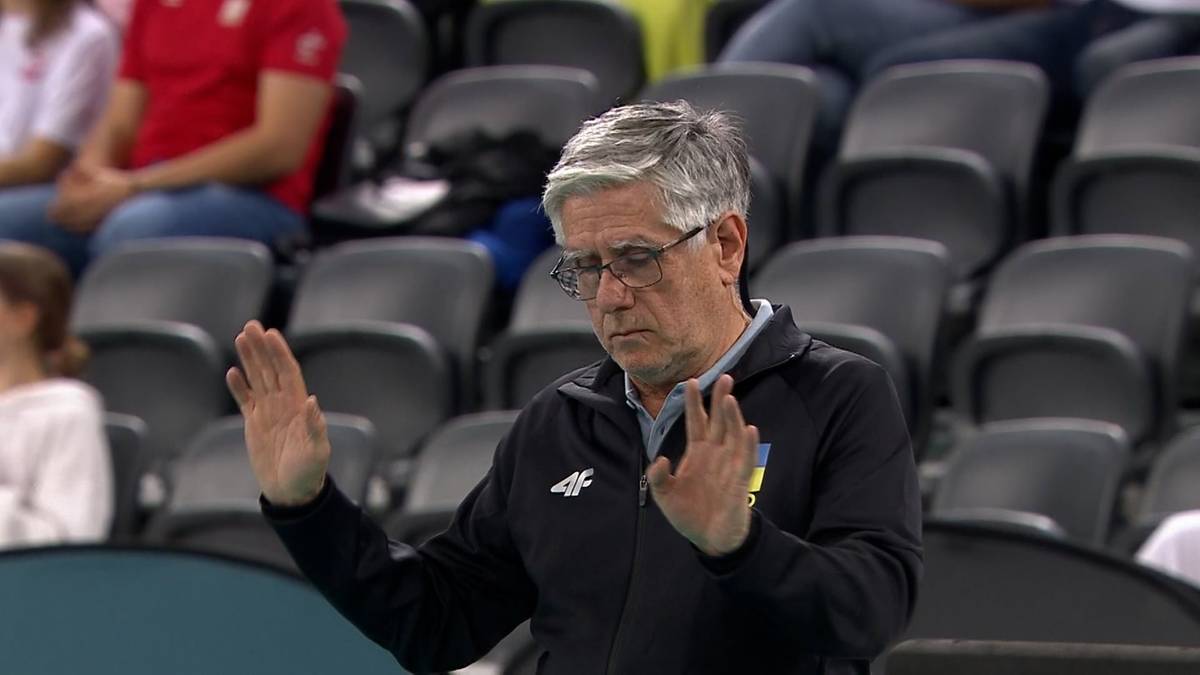 Mężczyzna w okularach, starszy, z siwymi włosami, ubrany w czarną bluzę z kapturem, z podniesionymi dłońmi, gestykuluje podczas meczu siatkówki.