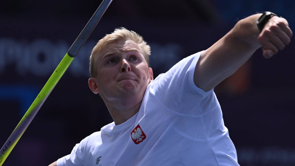 Młody mężczyzna o jasnych włosach w białej koszulce z polskim godłem wykonuje rzut oszczepem.