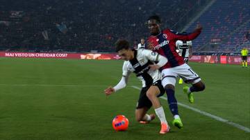 Dwóch piłkarzy walczy o piłkę na boisku, jeden w białej koszulce, drugi w czerwonej. Piłka znajduje się przed nimi.