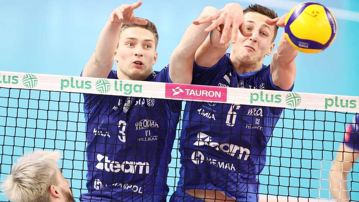 Dwóch siatkarzy w niebieskich strojach po tej samej stronie siatki podczas meczu. Gracz po lewej próbuje zablokować piłkę, a gracz po prawej wykonuje atak.