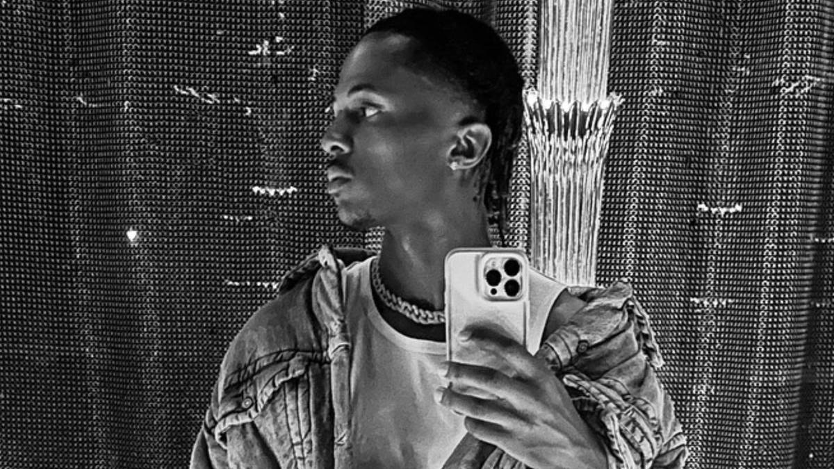 Młody mężczyzna z warkoczami, ubrany w T-shirt i kurtkę dżinsową, trzyma telefon kom&oacute;rkowy i robi sobie zdjęcie.