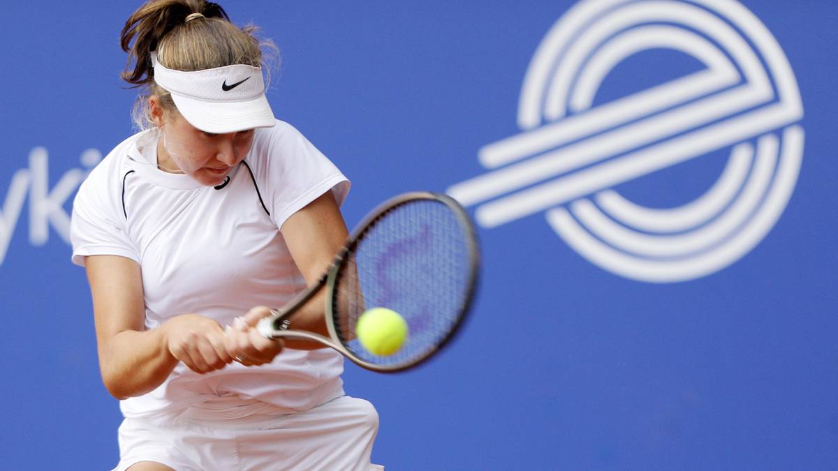 Tenisistka w białej koszulce i spódniczce, z daszkiem na głowie, odbija piłkę rakietą podczas meczu na kortach o niebieskim tle.