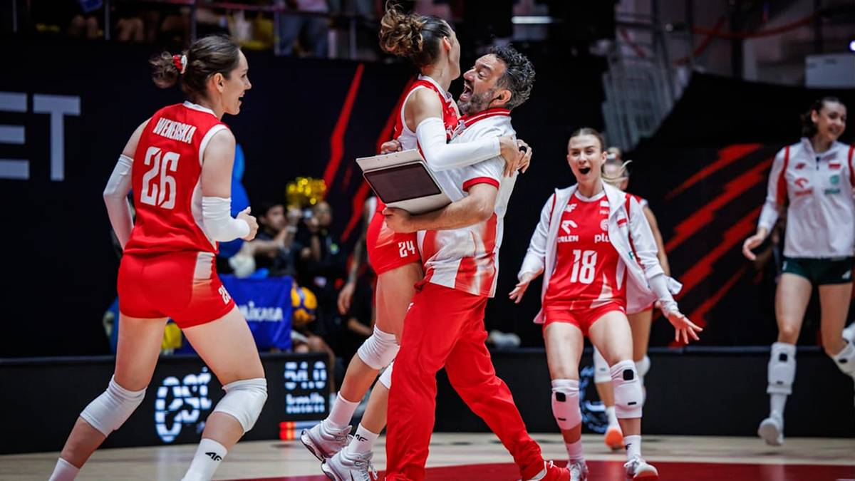 Trener obejmuje zawodniczkę siatkówki po wygranym meczu, oboje cieszą się ze zwycięstwa. Inne zawodniczki w tle również świętują.