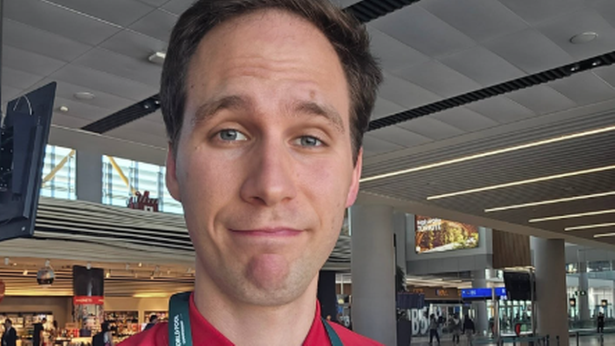 Młody mężczyzna z jasnymi włosami i niebieskimi oczami, ubrany w czerwoną koszulkę z widocznym fragmentem medalu, uśmiecha się do kamery. W tle widać jasne, nowoczesne wnętrze lotniska lub hali widowiskowej z dużą ilością światła naturalnego i sztucznego.