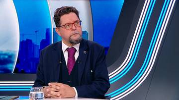 Mężczyzna w okularach i garniturze siedzi przy stole w studiu telewizyjnym.