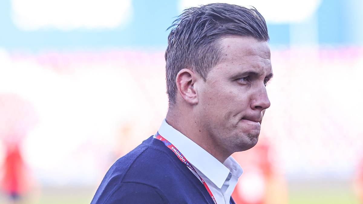 Portret mężczyzny w średnim wieku, ubranego w białą koszulę i granatowy sweter. Ma krótkie, ciemne włosy i spogląda w bok z zamyśloną miną. W tle widać rozmazane sylwetki ludzi i fragment trybun stadionu.