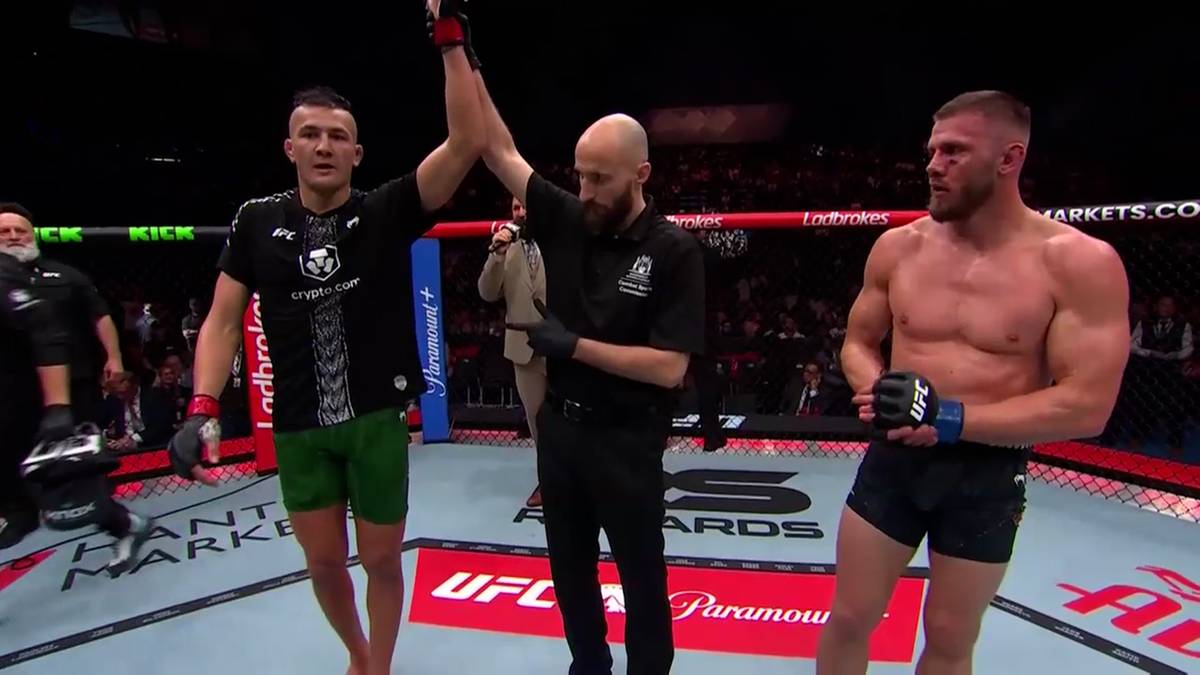 Dw&oacute;ch zawodnik&oacute;w MMA stoi w oktagonie, jeden z nich ma uniesioną rękę zwycięzcy, a sędzia stoi pomiędzy nimi.