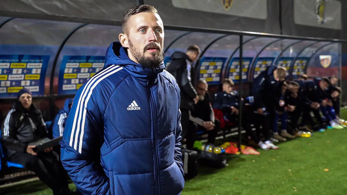 Trener w kurtce sportowej stoi na tle ławki rezerwowych z drużyną piłkarską.