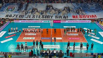 Widok z g&oacute;ry na boisko do siatk&oacute;wki podczas meczu PlusLigi, z zawodnikami na parkiecie i kibicami na trybunach.