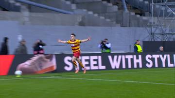 Piłkarz w ż&oacute;łto-czerwonym stroju świętuje strzelenie bramki, z rękami rozłożonymi na boki. Na tle stadionu widoczny jest duży ekran z napisem "STEEL WITH STYLE".