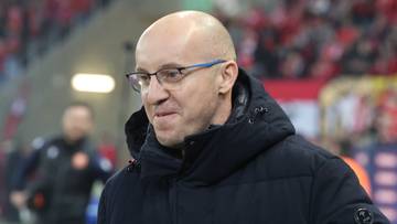 Trener w okularach i ciemnej kurtce na tle trybun stadionu z kibicami.