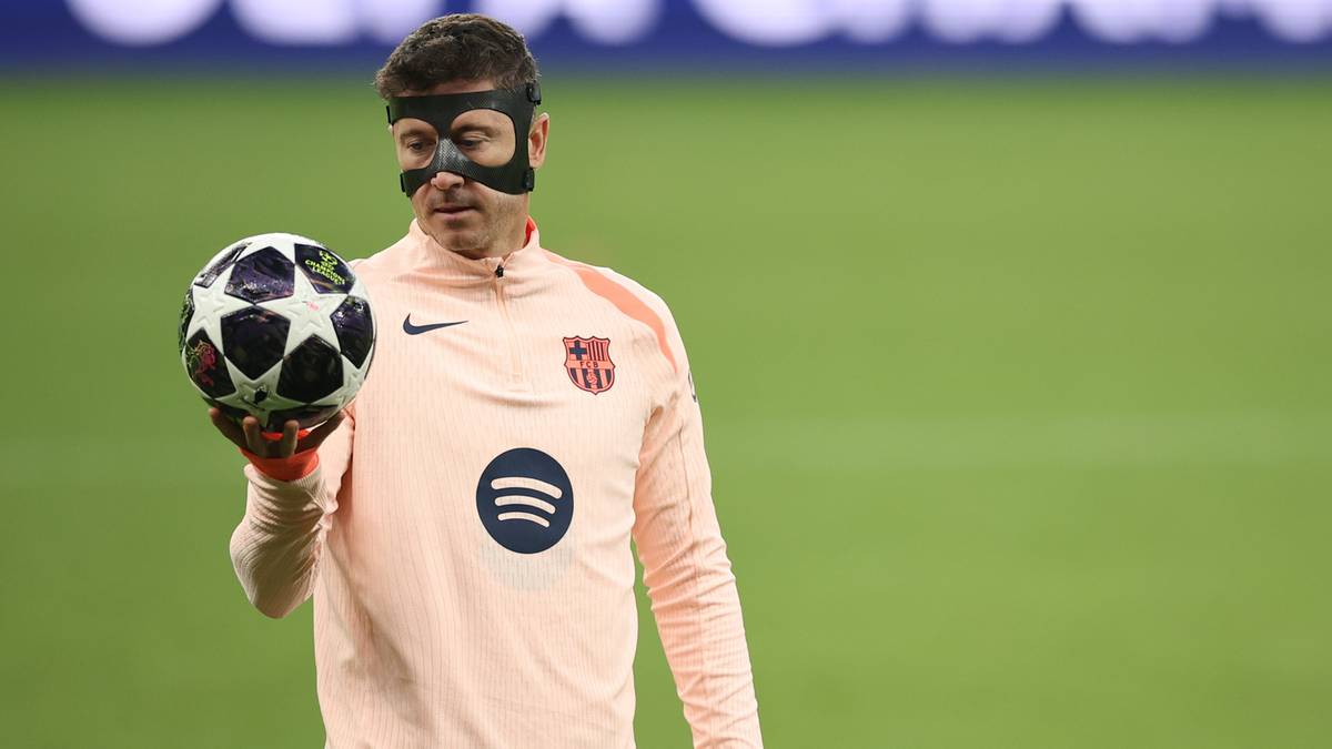 Piłkarz w r&oacute;żowej koszulce z długim rękawem i okularach ochronnych trzyma piłkę w lewej ręce na zielonym boisku.