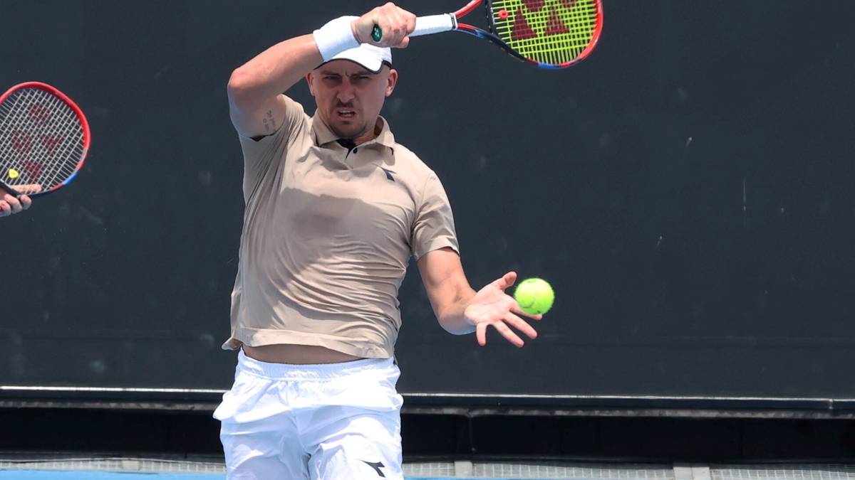 Dwóch tenisistów na korcie. Jeden zawodnik w beżowej koszulce i białych spodenkach, wykonuje zamach rakietą do uderzenia piłki. Drugi zawodnik, w niebieskim stroju, stoi w tle.