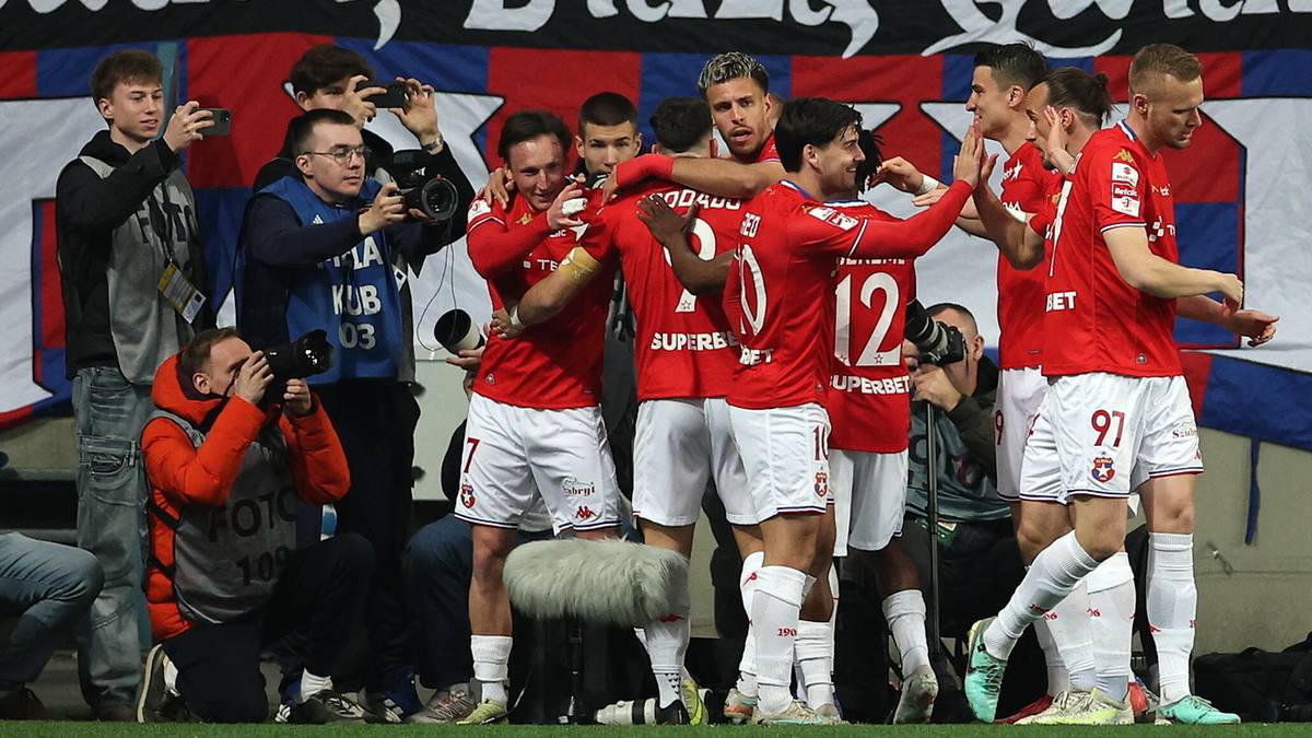 Piłkarze w czerwonych strojach z numerami na plecach świętujący na boisku piłkarskim, w tle baner z napisem "Biała Gwiazda".