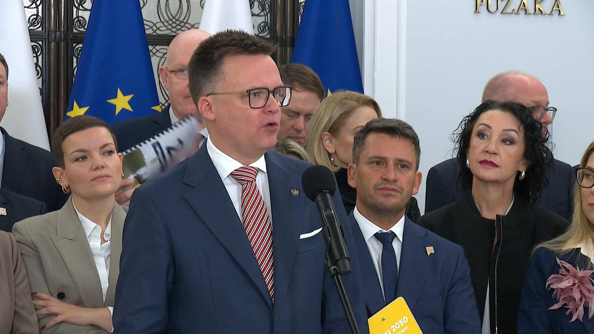 Mężczyzna w okularach i garniturze przemawia do mikrofonu, otoczony przez innych ludzi.