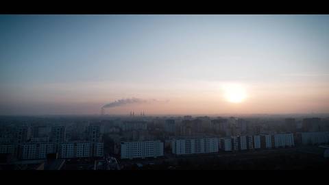 Panorama miasta o wschodzie lub zachodzie słońca, z widocznym słońcem, niebem w odcieniach błękitu i r&oacute;żu oraz dymem unoszącym się z fabrycznych komin&oacute;w w oddali.