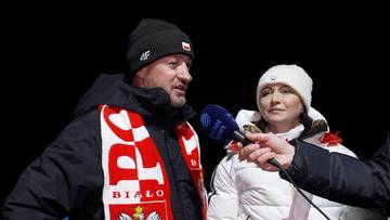 Adam Małysz w czapce i szaliku z polskimi barwami, udzielający wywiadu. Obok niego stoi kobieta w białej odzieży.